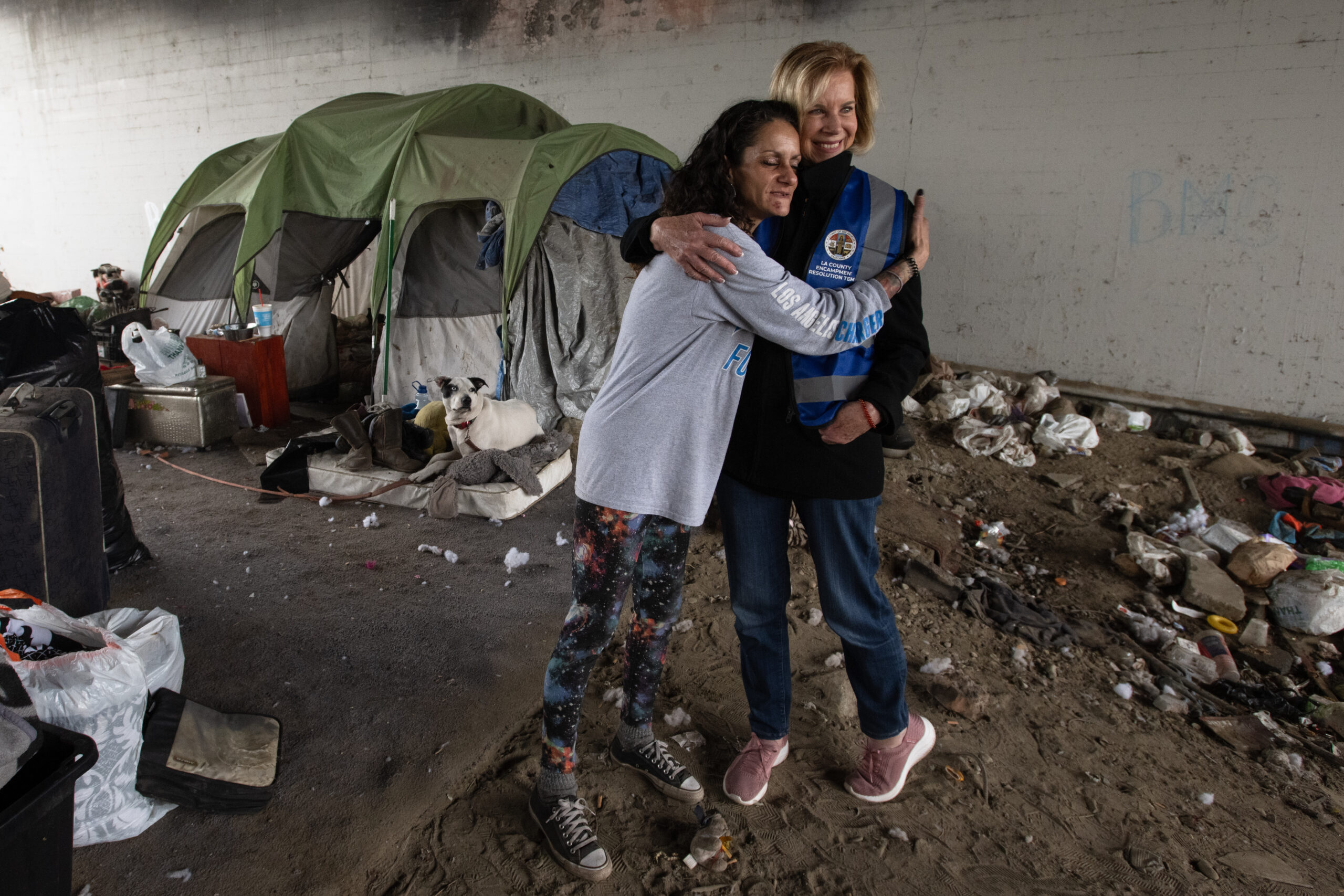 A woman wearing patterned leggings embraces Supervisor Hahn, who is wearing a blue safety vest under an overpass where several tents and belongings are set up. A dog rests on a mattress near the tents, and scattered trash and debris cover the ground around them.