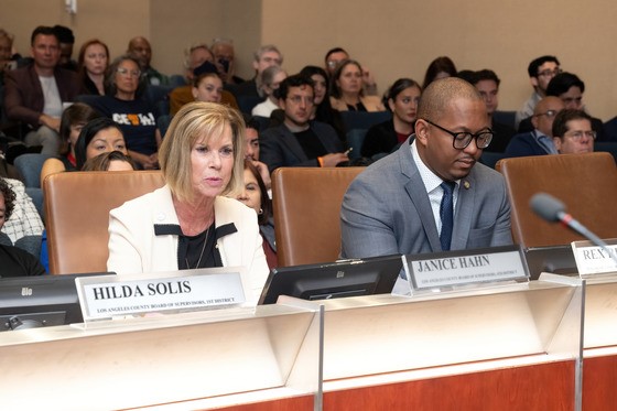 Supervisor Hahn in a light-colored blazer sits at a panel desk beside a man in a gray suit and blue tie; both face forward with nameplates in front of them, including one labeled 'Janice Hahn' and another labeled 'Hilda Solis' partially visible; microphones and tablets sit on the desk, and a large audience fills the rows behind them, watching attentively. 