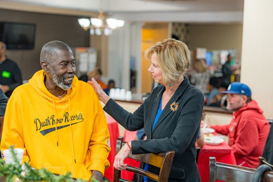 A woman in a dark blazer gently rests her hand on the shoulder of an older man wearing a bright yellow “Death Row Records” hoodie. They are smiling and talking inside a busy community center or dining hall, where other people sit at tables covered with red tablecloths. Warm lighting and holiday greenery create a welcoming atmosphere.