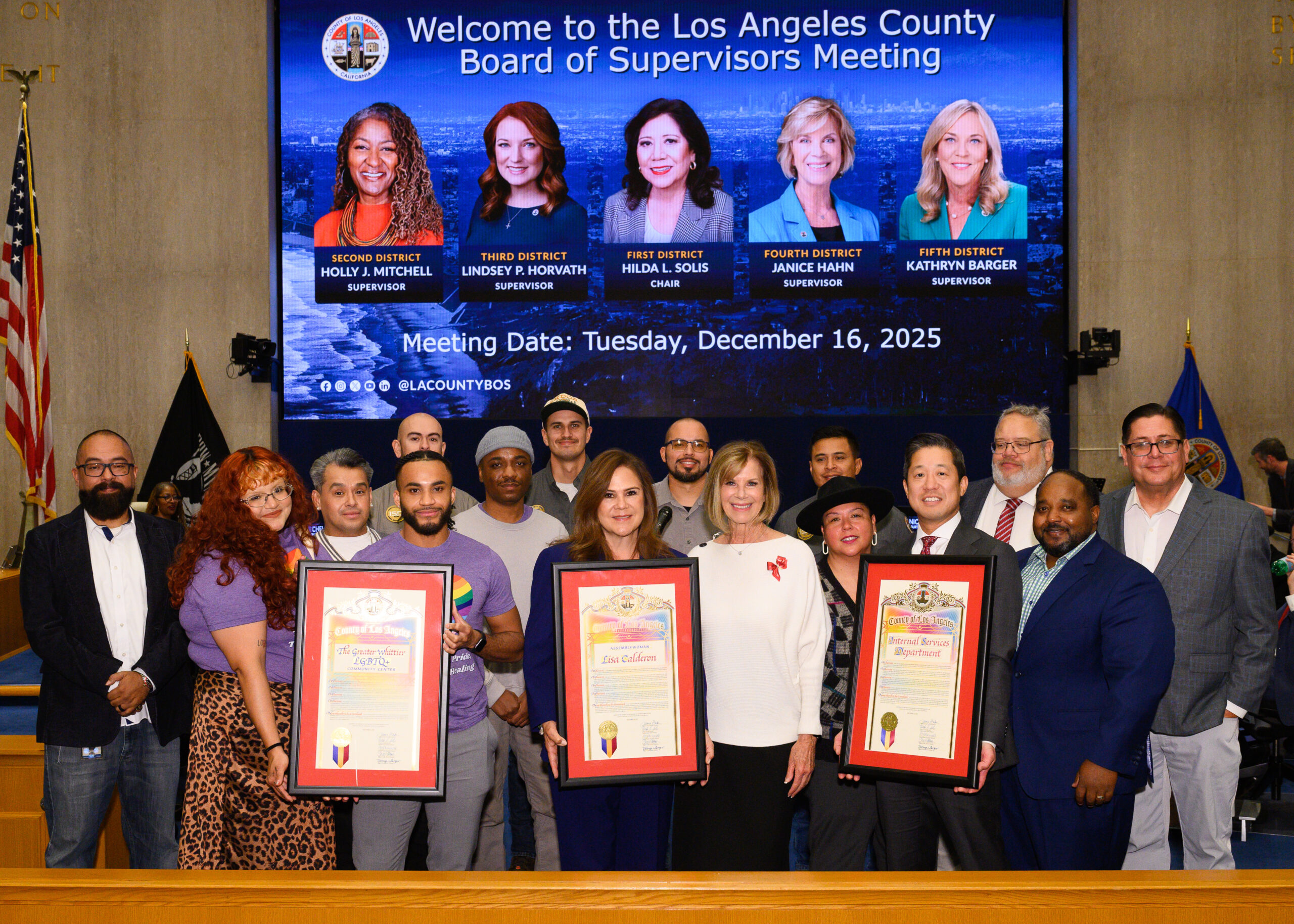 A large group of people pose inside a government meeting chamber, standing behind a wooden dais. Several people in the front hold framed proclamations. Behind them, a large screen reads &ldquo;Welcome to the Los Angeles County Board of Supervisors Meeting&rdquo; with portraits of the five supervisors and the meeting date, Tuesday, December 16, 2025. American and county flags are visible on either side, suggesting a formal civic recognition or award presentation.