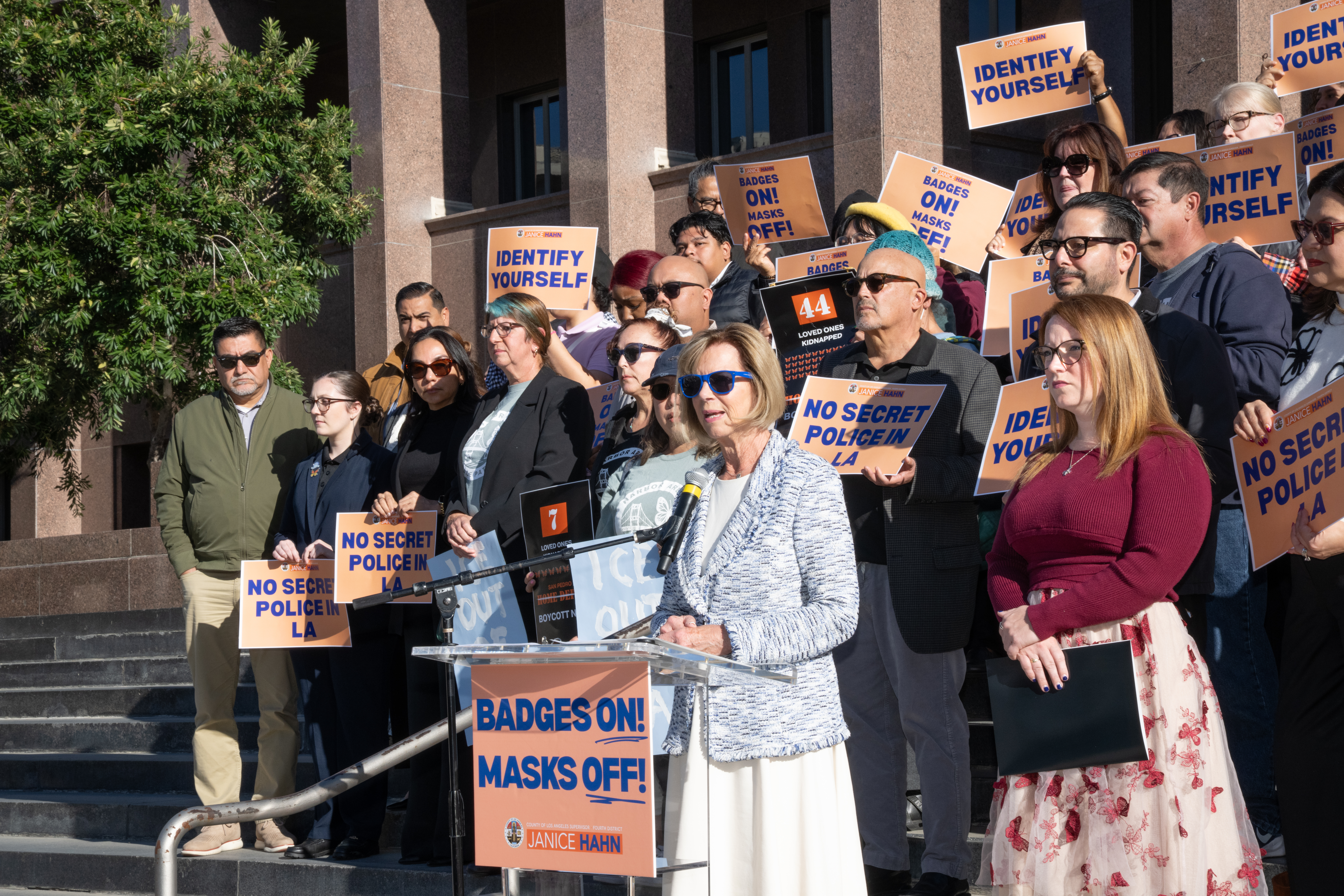 A woman speaks at a podium on the steps of a government building, surrounded by a large group of people holding signs that read “Badges On! Masks Off!,” “No Secret Police in LA,” and “Identify Yourself.” The crowd stands closely together behind her, many wearing sunglasses, while listening to her remarks. A tree and the building’s stone façade are visible in the background.