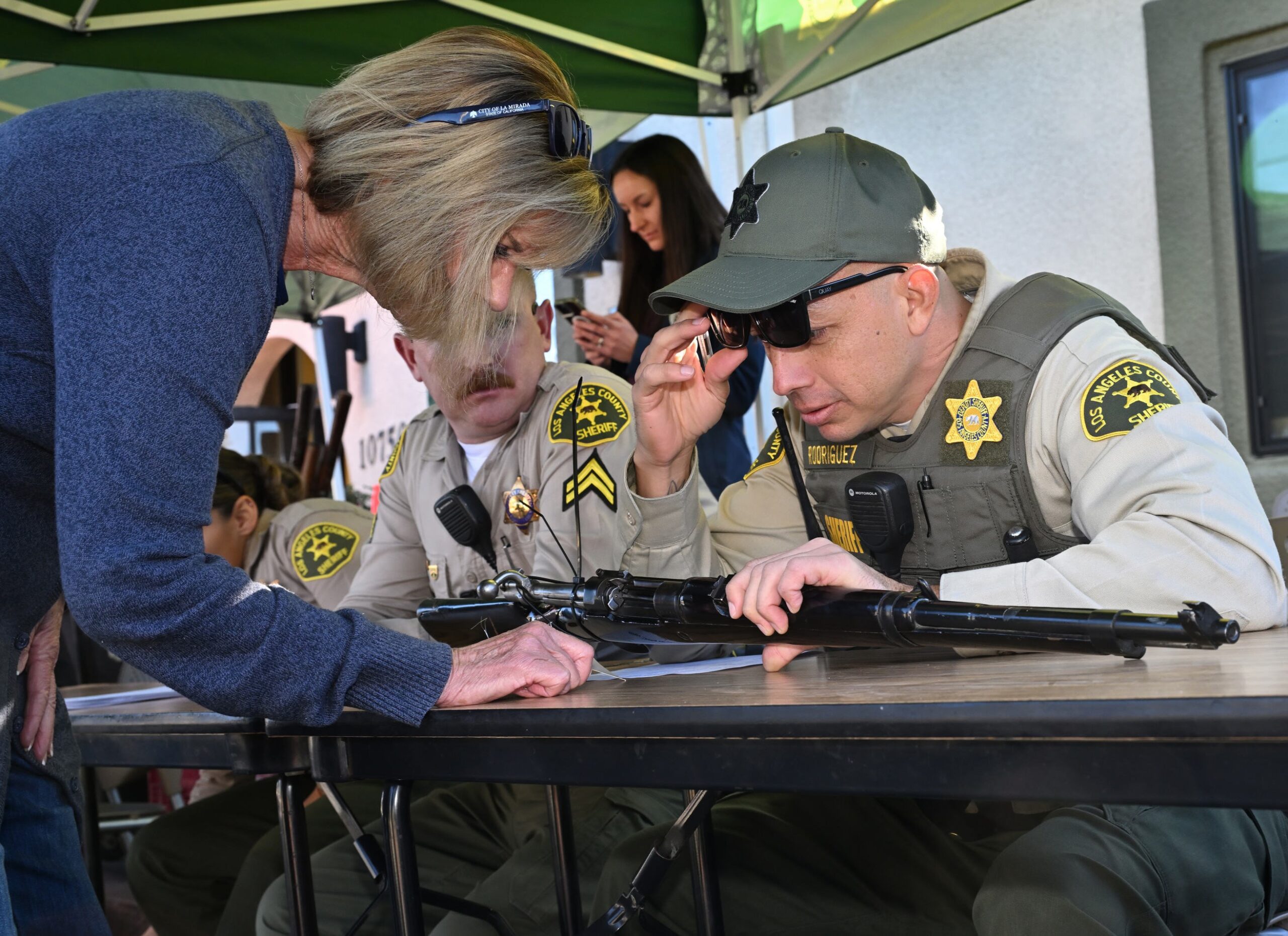 A woman leans over a table to speak with two uniformed sheriff&rsquo;s deputies seated under a canopy. One deputy is adjusting his sunglasses while examining a long firearm laid on the table, and the other deputy watches attentively. A third person stands in the background using a phone.