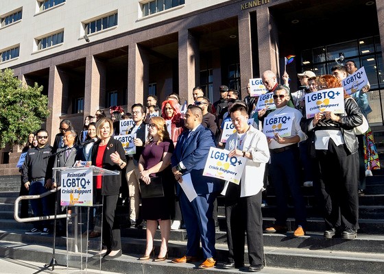 A large group of people holding signs that say "LGBTQ+ Crisis Support" stand on steps outside of a building as a woman speaks at a podium.