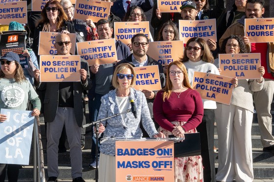 A woman speaks at a podium on the steps of a public building, accompanied by another woman standing beside her. Behind them, a large group of people hold orange and blue signs that read “Identify Yourself,” “Badges On! Masks Off!,” and “No Secret Police in LA.” The crowd stands closely together, facing forward in support of the message.
