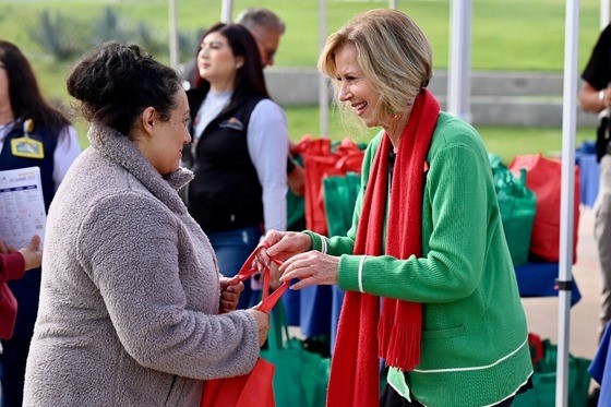 A woman wearing a green sweater and red scarf hands a red tote bag to another woman who is wearing a grey sweater.