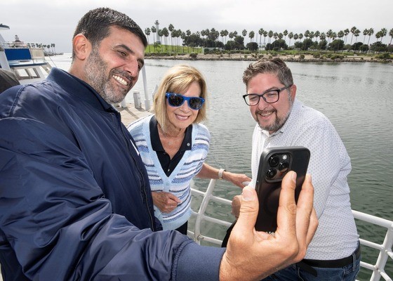 Three adults stand on a dock beside calm water, smiling as one of them holds up a smartphone to take a selfie. Palm trees line the shore in the background, and a boat is partially visible to the left. The group appears cheerful, leaning in close together.