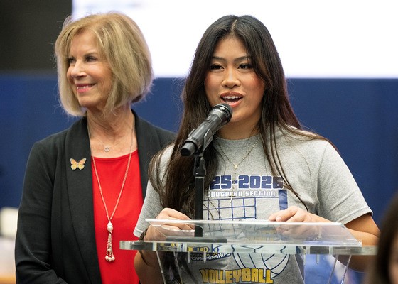 A young woman stands at a clear podium speaking into a microphone, wearing a gray volleyball championship T-shirt, while an older woman stands slightly behind her smiling. The background appears to be an indoor event or ceremony space.