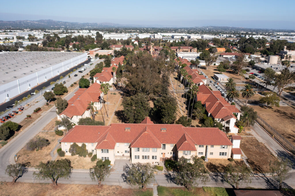 Aerial photograph of a campus-like residential complex with several two-story cream-colored buildings topped with red tile roofs. The buildings are arranged around a dry, tree-filled central area with tall palm trees and walking paths. A narrow street with parked cars runs along the right side, and large industrial warehouse buildings stretch across the background, with low mountains visible in the distance.