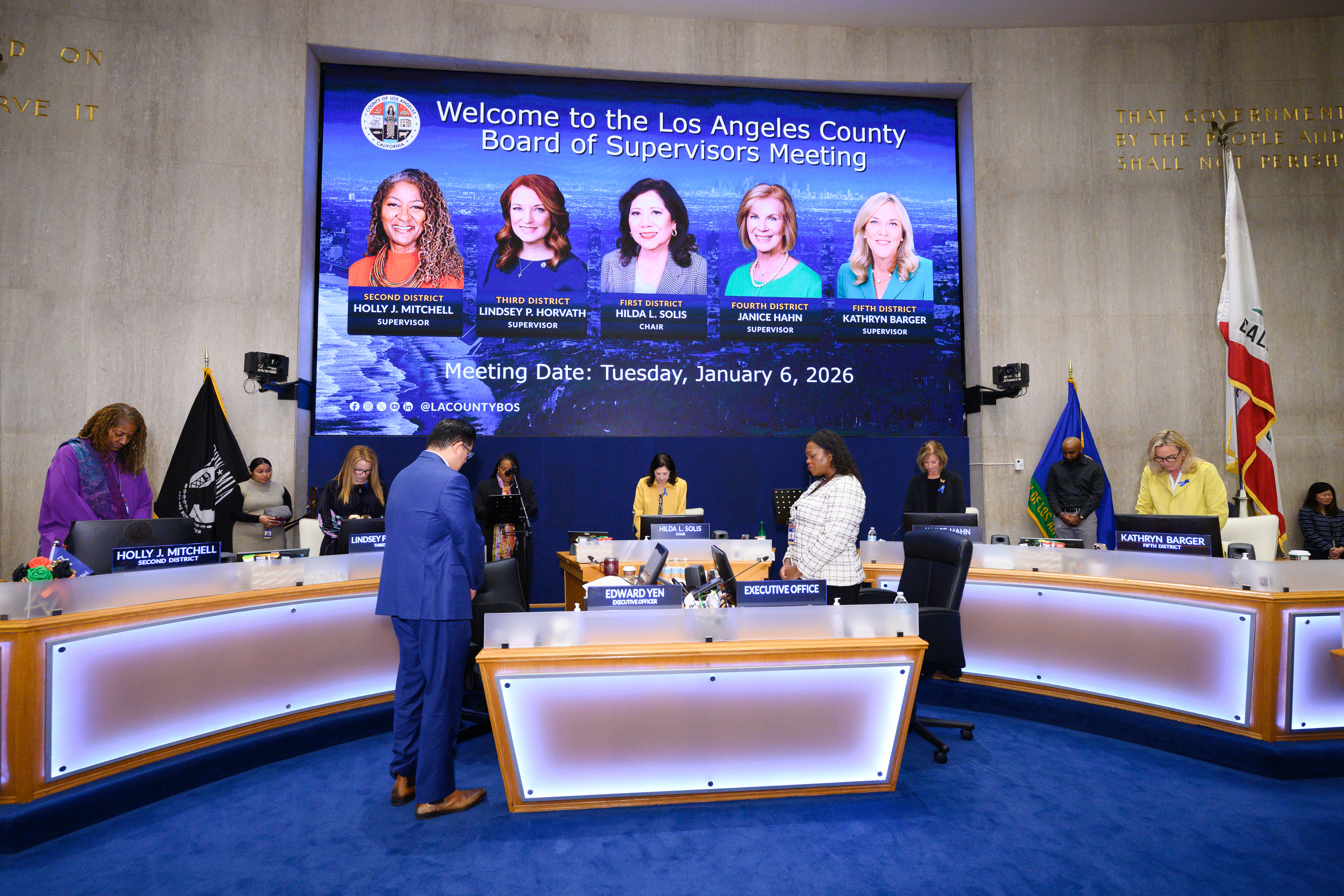 A formal meeting room for the Los Angeles County Board of Supervisors, with officials standing at a curved dais during what appears to be a moment of silence. A large digital screen behind them displays &ldquo;Welcome to the Los Angeles County Board of Supervisors Meeting&rdquo; along with headshots and names of the five supervisors and the meeting date, January 6, 2026. U.S. and California flags stand to the right, and staff members and attendees are positioned around the room.