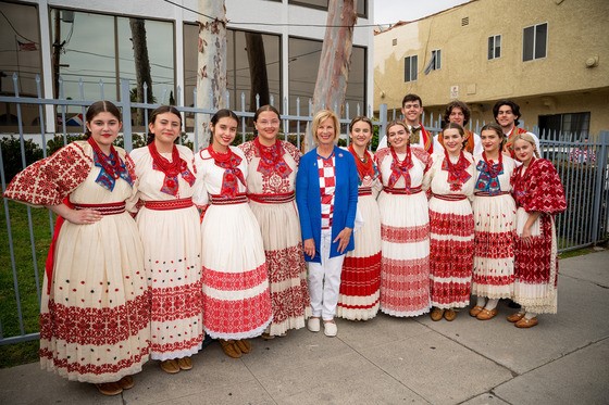 Supervisor Janice Hahn stands with a group of people outdoors on a sidewalk, smiling at the camera. The group is wearing coordinated traditional-style outfits in red, white, and cream, with embroidered patterns and matching scarves, while Supervisor Hahn is in the center wearing a blue blazer over a patterned blouse and white pants. A metal fence, trees, and residential buildings appear in the background.