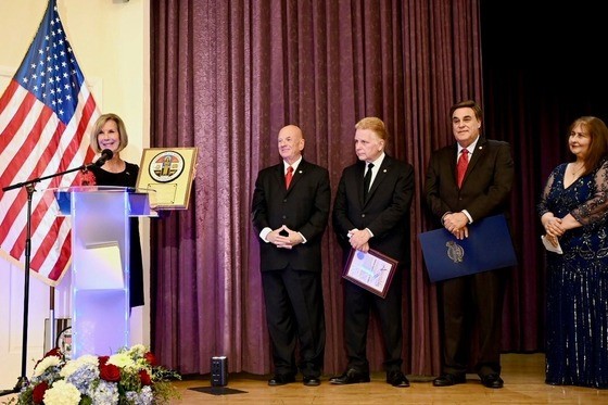 A formal ceremony on a stage with a woman speaking at a podium bearing a city seal, with a U.S. flag displayed beside her. Four adults stand to her right, dressed in formal attire; two men hold framed certificates or folders. A curtain backdrop and floral arrangement at the front of the stage complete the setting.