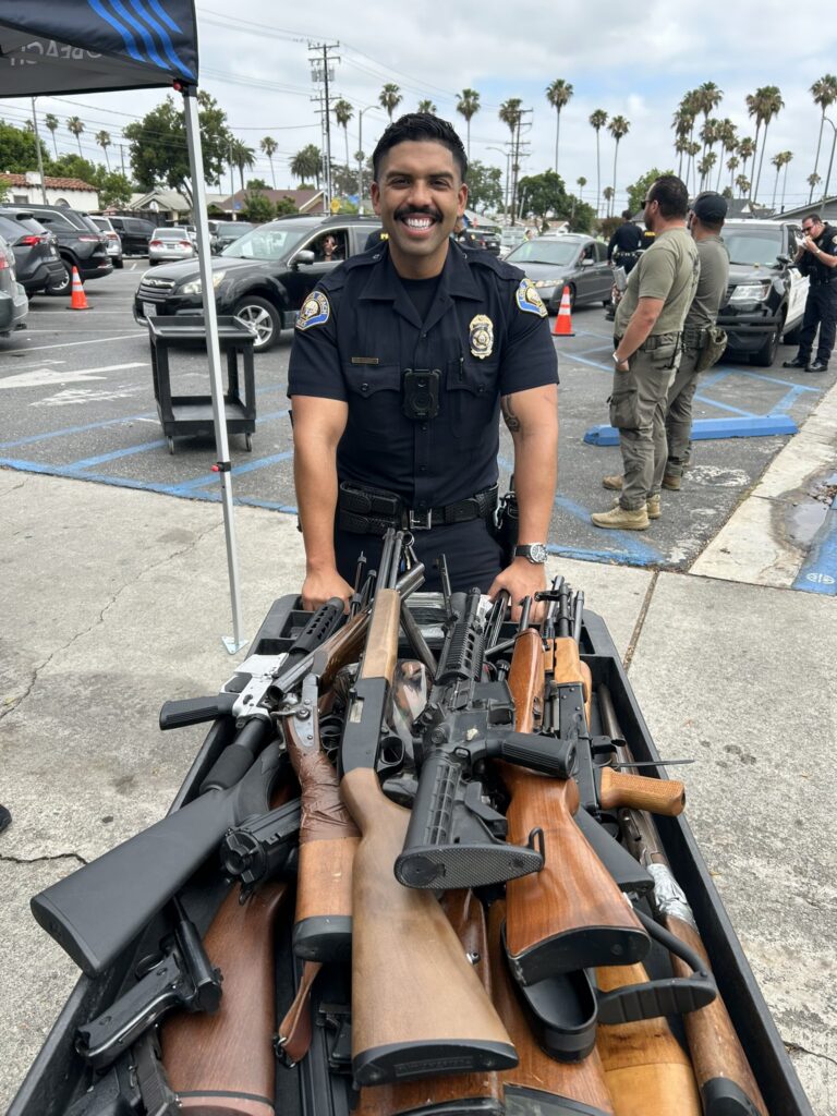 A smiling uniformed police officer stands behind a rolling cart filled with a variety of firearms, including rifles, shotguns, and handguns, at an outdoor community event in a parking lot. The officer is wearing a body camera and badge, while several other law enforcement personnel, parked vehicles, orange traffic cones, and tall palm trees are visible in the background under a cloudy sky.